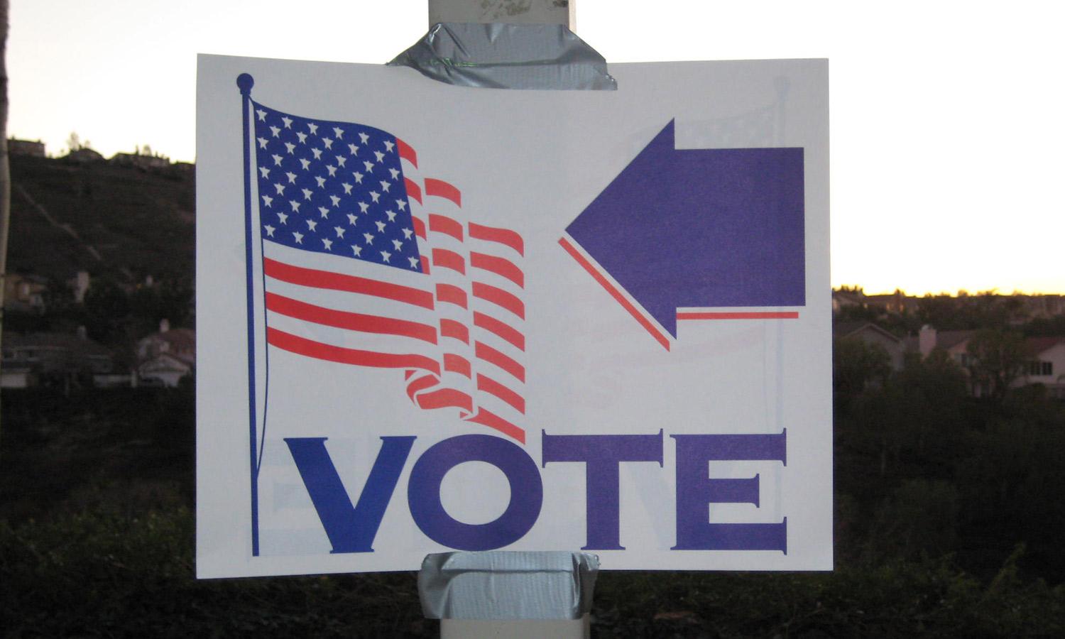 A voting sign on a lamppost with American flag