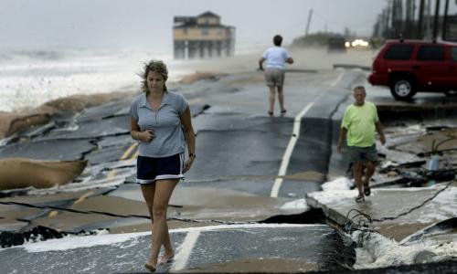 Three people walk on a road that's been broken apart by a storm. 
