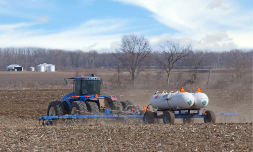 photo of a tractor in a turned-over field pulling a trailer with two tanks of anhydrous ammonia fertilizer that is being sprayed on the field; farm buildings and silos are in the distance